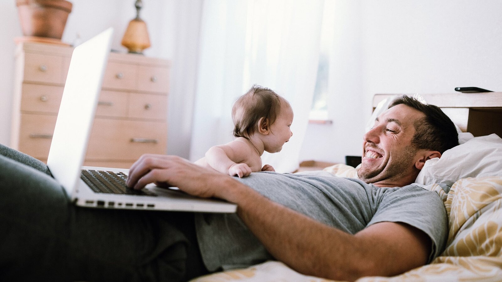 Vater liegt lachend mit Kleinkind im Bett und verwaltet bequem seinen Stromtarif online