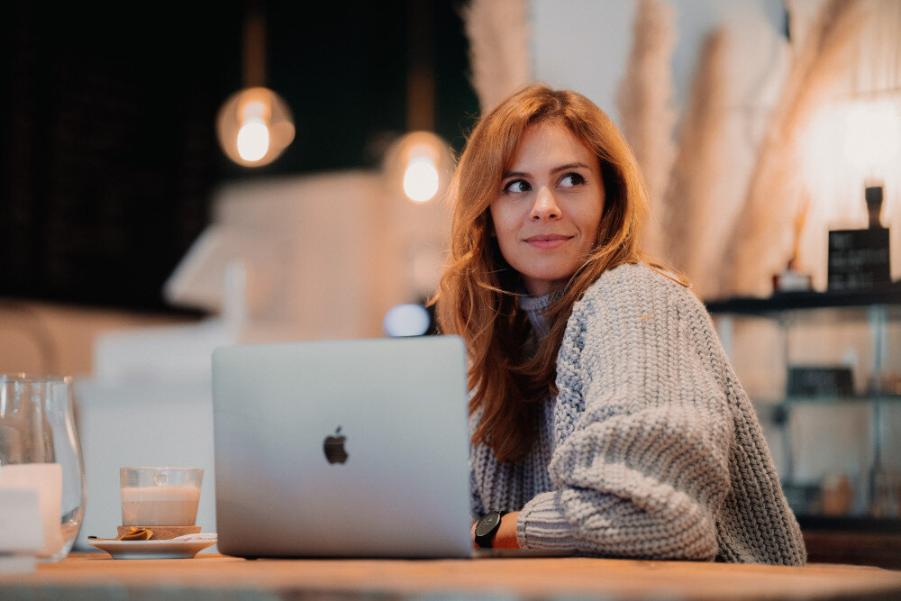 Eine junge rothaarige Frau in grauem Strickpullover sitzt an einem Holztisch vor ihrem Laptop und schaut in die Ferne.  | © jenny-ueberberg-eV2cVxPQ1tY-unsplash