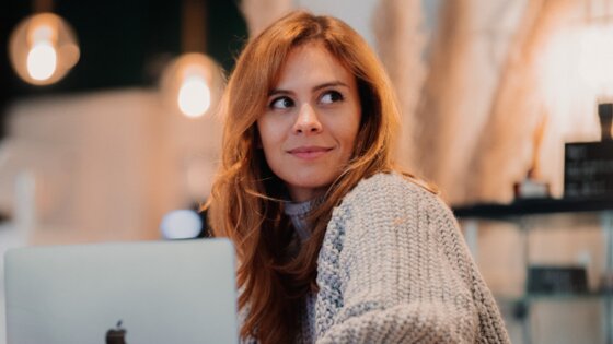Eine junge rothaarige Frau in grauem Strickpullover sitzt an einem Holztisch vor ihrem Laptop und schaut in die Ferne.  | © jenny-ueberberg-eV2cVxPQ1tY-unsplash