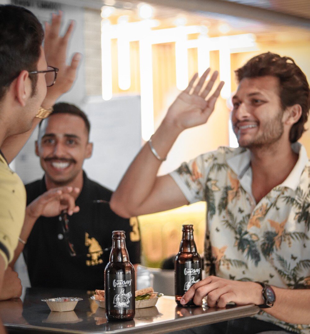 3 junge Männer sitzen in einer Bar, schlagen sich ein High-Five ein und trinken gesellig ein Bier zusammen. | © dollar-gill-EdnRBeOytlE-unsplash