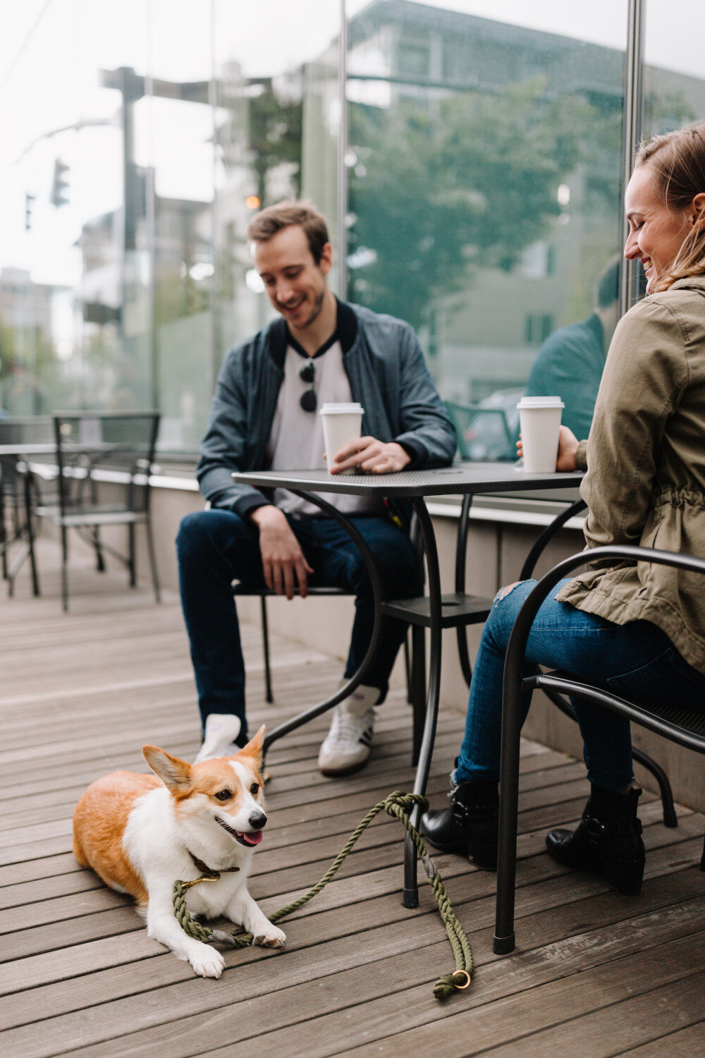 Eine junge Frau und ein junger Mann sitzen in einem Café im Sitzbereich draußen und lächeln. Ein Hund liegt auf dem Boden und relaxt. | © chewy-AQ8XIIRDuWc-unsplash