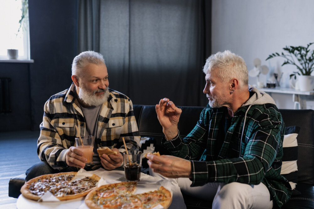 Zwei befreundete, Holzfällerhemden tragende Mid-Ager Freunde essen gemeinsam Pizza im Wohnzimmer. Einer von beiden signalisiert, dass sie prima schmeckt. | © pexels-alena-darmel-7322108