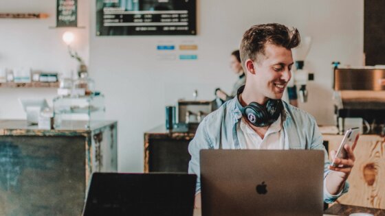 Junger Mann steht an einem Tresen in einem trendy Café, schaut auf sein Handydisplay und hat einen aufgeklappten Laptop vor sich. Um den Hals trägt er große Kopfhörer. | © austin-distel-TluMvvrZ57g-unsplash