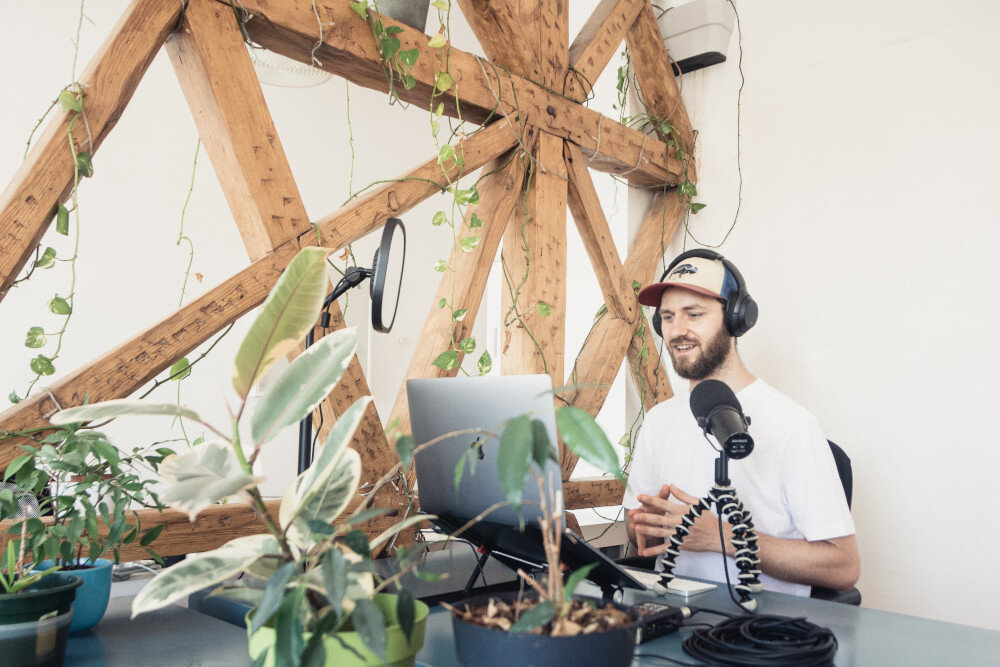 Ein junger Mann mit Cappy sitzt in seinem hell eingerichteten Podcast-Studio und steckt mitten in der Produktion | © jeremy-enns-1vrWiQvtxW8-unsplash