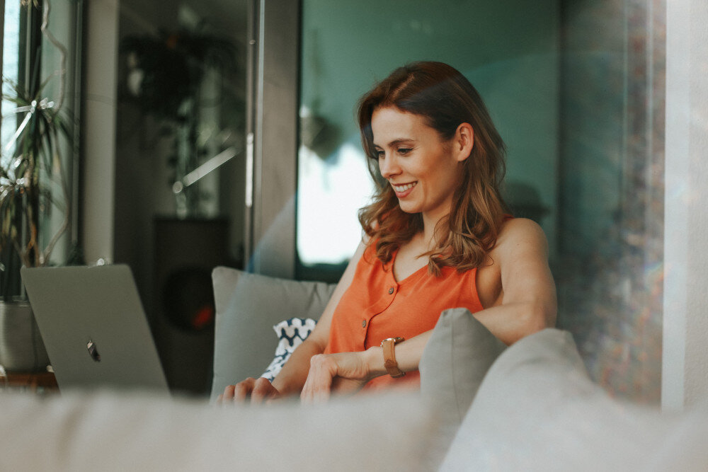 Eine Frau mit hellbraunem Haar und orangefarbenem Top sitzt auf Loungemöbeln in ihrer Wohnung und arbeitet in gemütlichem Umfeld an einem Laptop. Sie lächelt in die Kamera | © jenny-ueberberg-BaSeK7rwc1A-unsplash