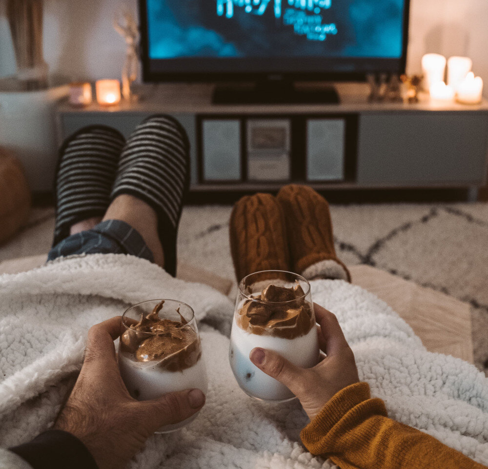 Ein Paar sitzt mit warmen Hausschuhen und zugedeckt auf der Couch. Es wird ein Heißgetränk mit Milch und Schokocreme in den Händen gehalten. Im TV läuft ein Film und auf dem Lowboard sind Kerzen angezündet. | © pexels-taryn-elliott-4488194-min