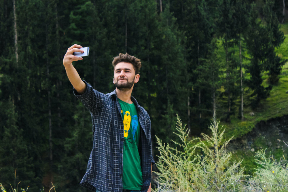Ein junger Mann in kariertem Hemd fotografiert sich selbst mit seinem Mobiltelefon im Wald.  | © pexels-farhan-ullah-baig-1053845