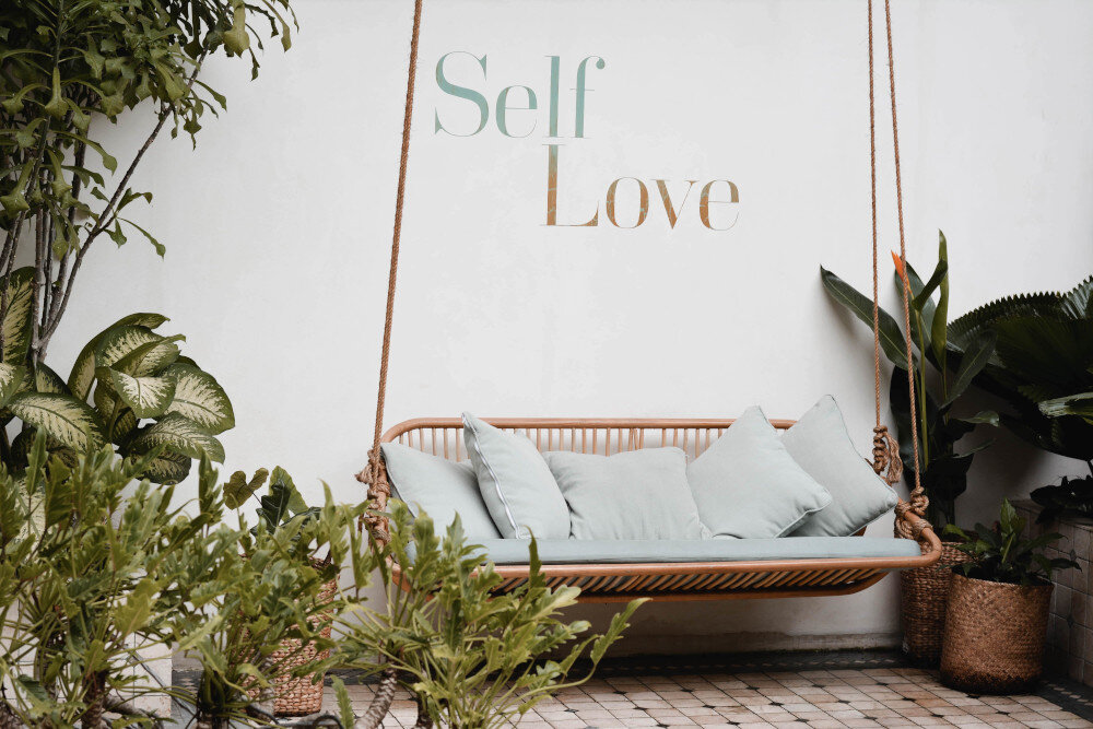 Es ist eine Hängeschaukel vor einer Wand zu sehen, die mit einer Wandbemalung mit dem Text "Self Love" verziert ist. Außenherum sieht man Grünpflanzen.  | © content-pixie-2V5PJgGqyts-unsplash