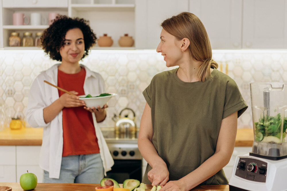 Ein junges gleichgeschlechtliches Damenpaar kocht gemeinsam in der Küche, beide Frauen lächeln sich vertraut an. | © pexels-mikhail-nilov-6707697
