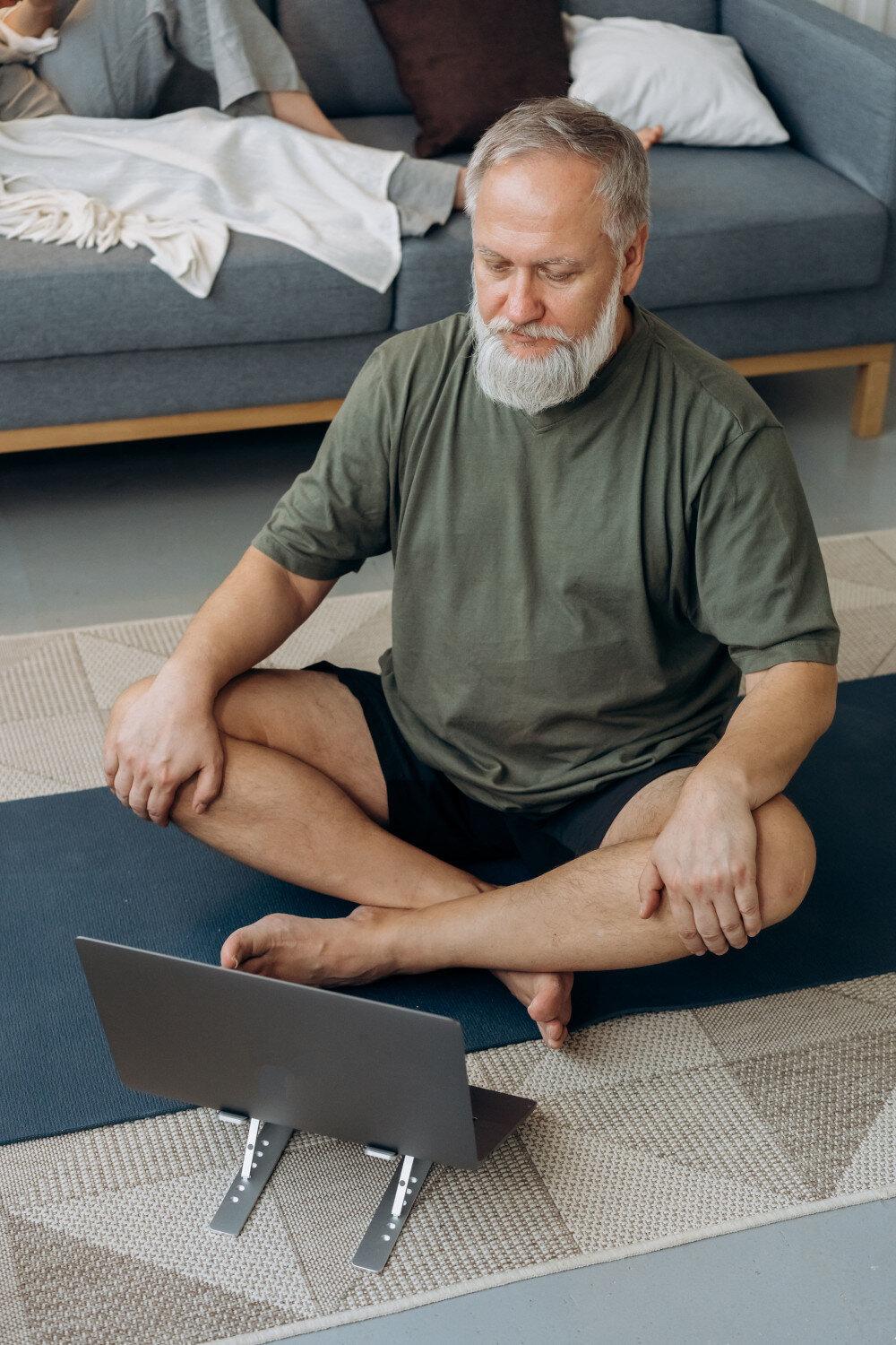 Ein Mann mit grauem Bart und Haupthaar sitzt auf einer Yogamatte auf dem Wohnzimmerboden und schaut sich ein Tutorial auf dem vor sich aufgebauten Tablet an- | © pexels-mikhail-nilov-7529994