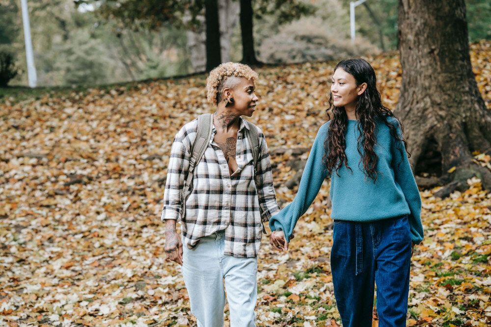 Ein gleichgeschlechtliches Damenpärchen läuft händchenhaltend durch einen Park, in dem viel herabgefallenes Herbstlaub liegt. | © pexels-tim-samuel-5845047