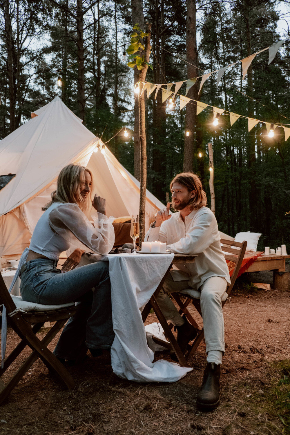 Junges Paar sitzt in der Natur an einem Holztisch vor einem Zelt und lacht. Sie genießen die romantische Abendstimmung. Es ist eine Lichterkette zum Zelt gespannt. | © pexels-cottonbro-studio-5365205