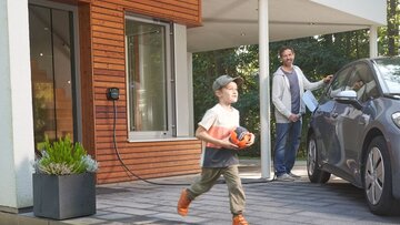 Familie nutzt umweltfreundliche Energie: Vater lädt E-Auto, Sohn spielt draußen im Garten