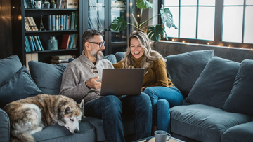 Zwei Personen mit Hund auf der Couch, lachend vor dem Laptop