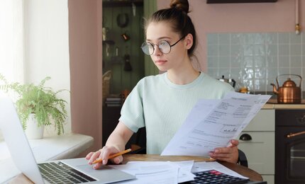 Junge Frau sitzt vor Laptop, schaut auf Dokumente und Rechnungen