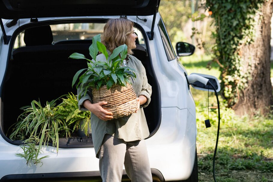 Frau mit grüner Topfpflanze steht neben einem ladenden E-Auto