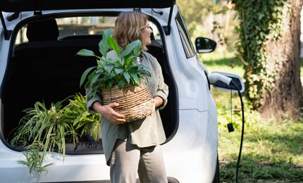 Frau mit grüner Topfpflanze steht neben einem ladenden E-Auto
