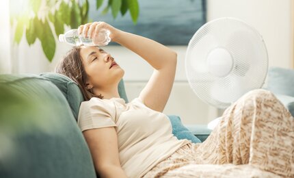 Frau bei heißem Wetter auf der Couch mit Ventilator und Wasserflasche